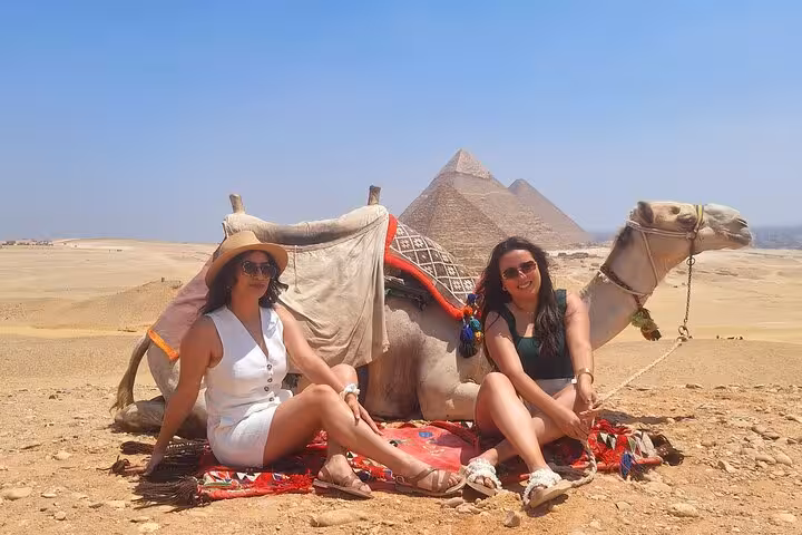 Two tourists sitting with a camel in front of the Pyramids of Giza, enjoying a scenic Egyptian tour experience.