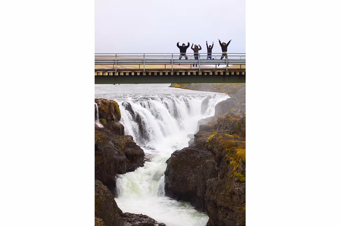 Four excited tourists jumping on a bridge over a scenic waterfall canyon in North Iceland