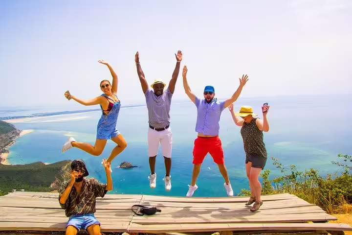 Group of tourists jumping joyfully on a scenic cliffside in Arrábida, Portugal, overlooking the turquoise ocean and lush landscape.