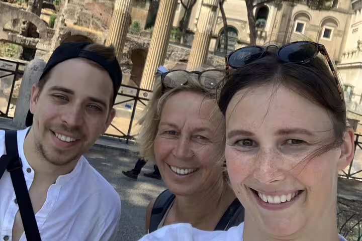 Three smiling tourists taking a selfie at ancient Roman ruins with stone columns in Rome