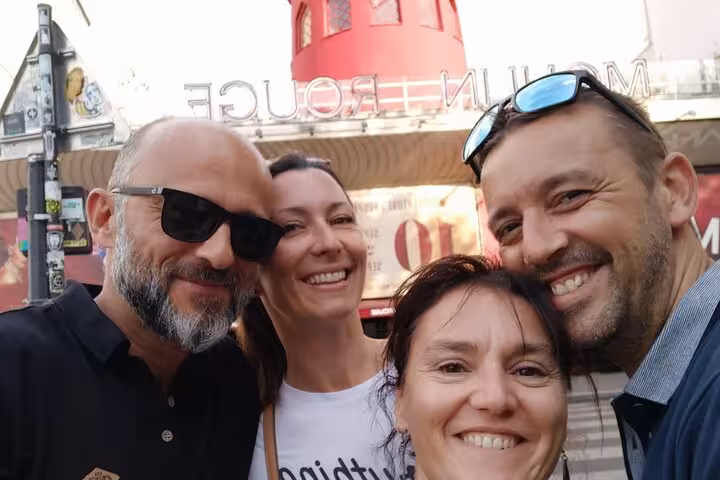 Group of four smiling tourists taking a selfie in front of the Moulin Rouge in Paris