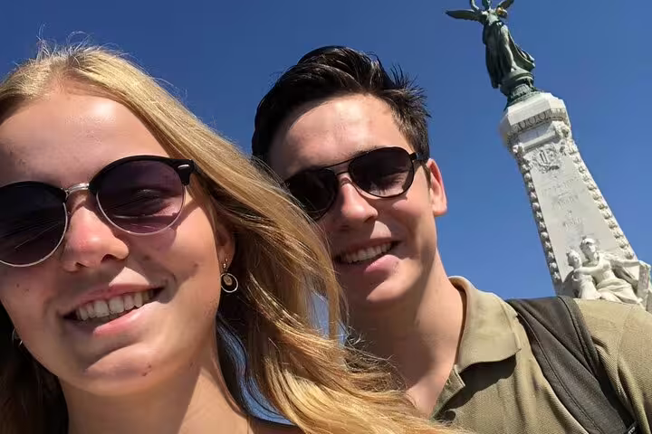 Two smiling tourists in sunglasses taking a selfie near a monument in Nice on a sunny day