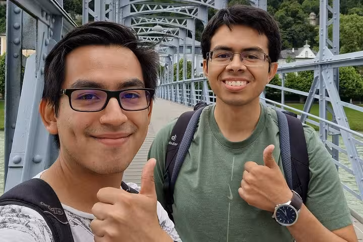 Two smiling young men giving thumbs up on a historic iron bridge in Salzburg Old Town