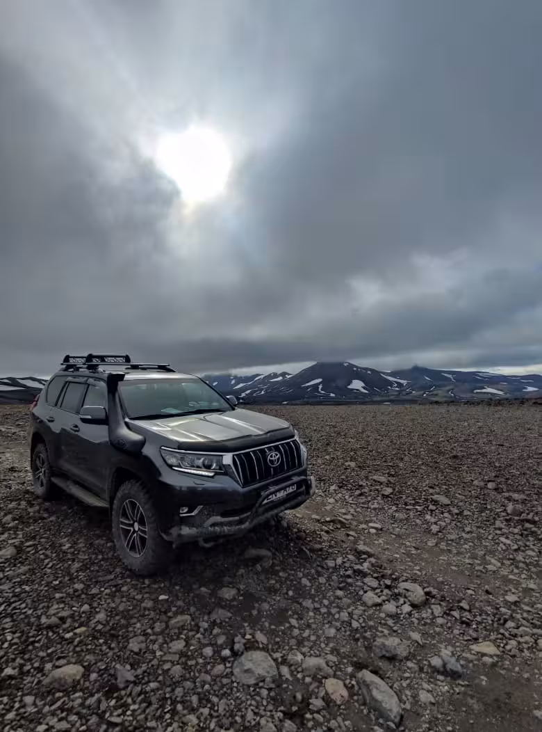 Black Toyota Land Cruiser Prado on rocky Iceland highland terrain with snow-capped volcanic mountains under dramatic cloudy sky