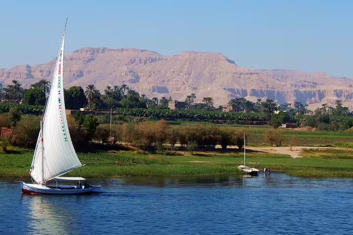 Traditional felucca sailboat on the Nile in Aswan, Egypt with palm-lined shore and desert hills on a sailing ride