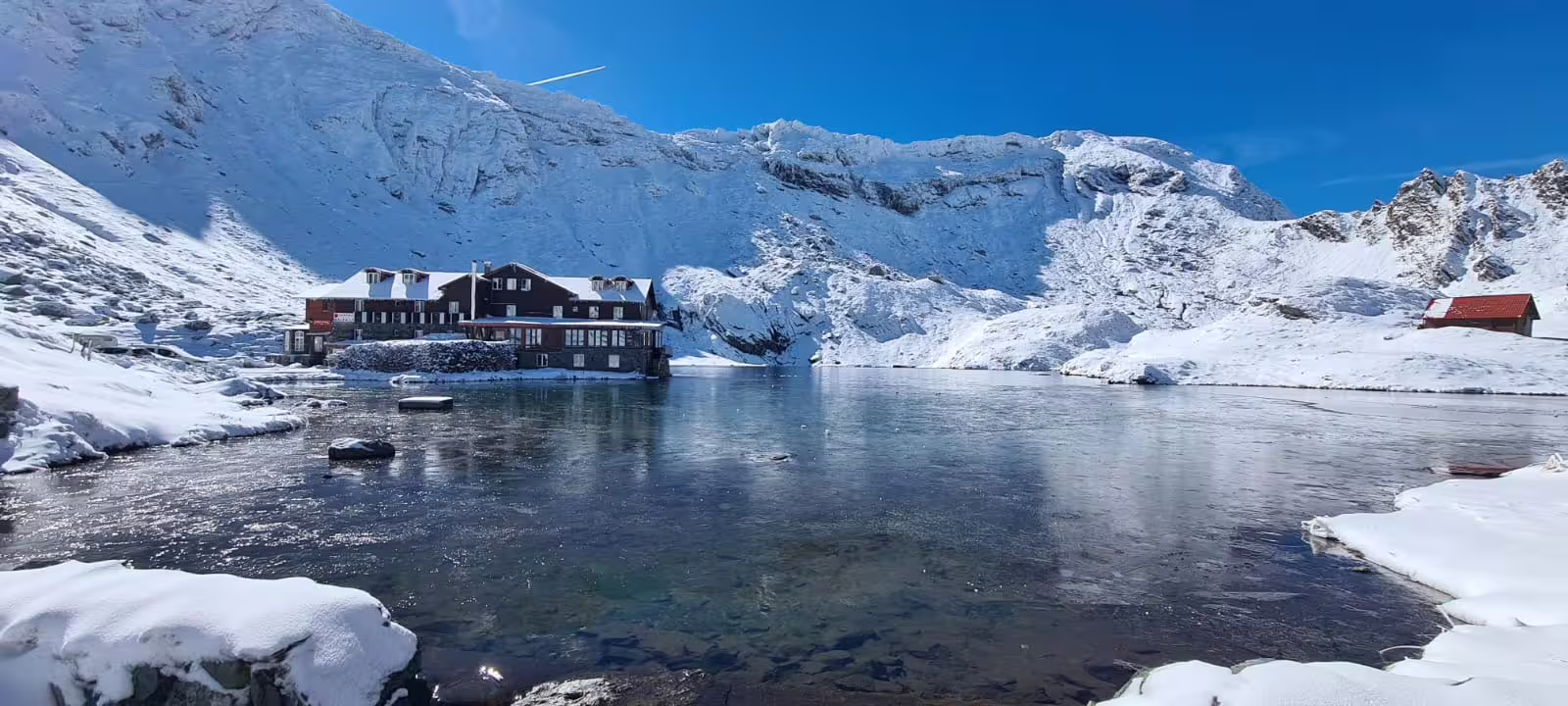 Frozen alpine lake beside a mountain chalet surrounded by snow-covered peaks on Transfagarasan Road