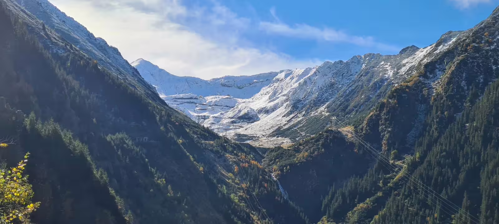 Snow-capped Transfagarasan mountain valley with forested slopes and power lines under blue sky
