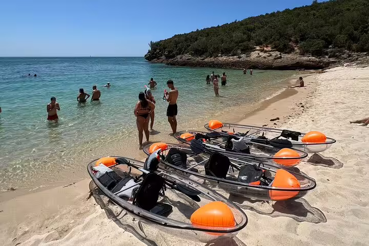 Transparent kayaks lined up on a sandy beach in Arrábida, with people enjoying the clear blue sea in the background.