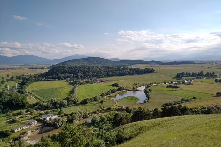 Aerial view of Transylvania valley with green fields, a small pond, forested hills, and mountains in the background