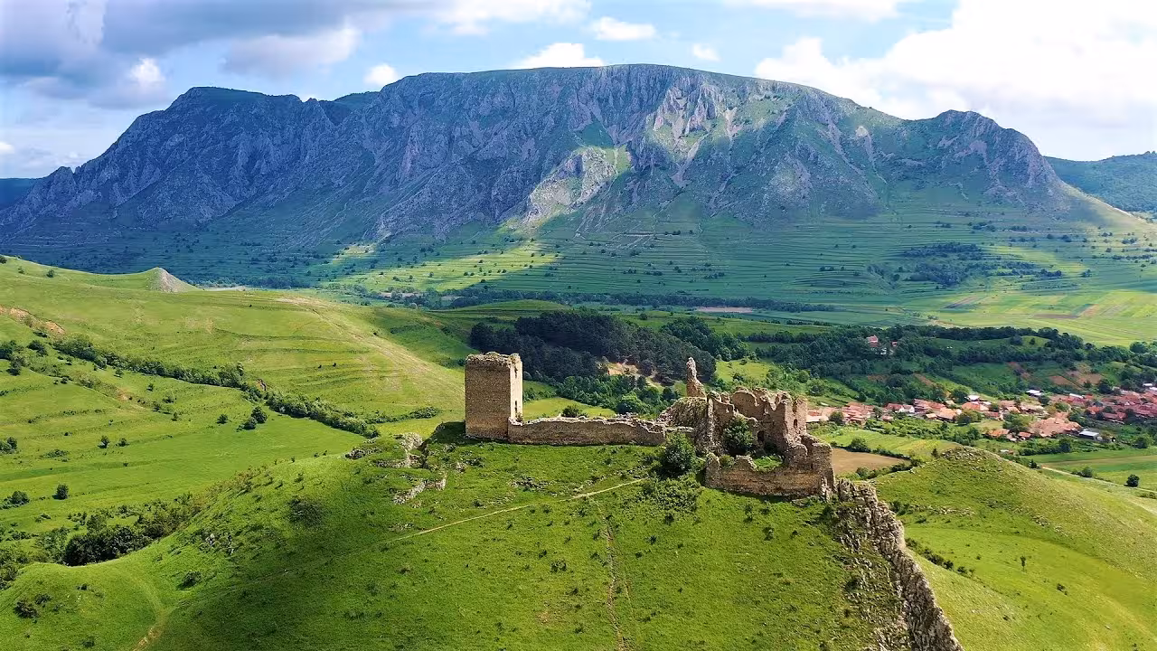 Medieval castle ruins on a hilltop with lush green valleys and dramatic mountain backdrop in Transylvania