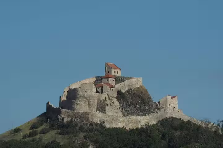 Medieval hilltop castle ruins with stone walls and red-roofed buildings under a clear blue sky in Transylvania