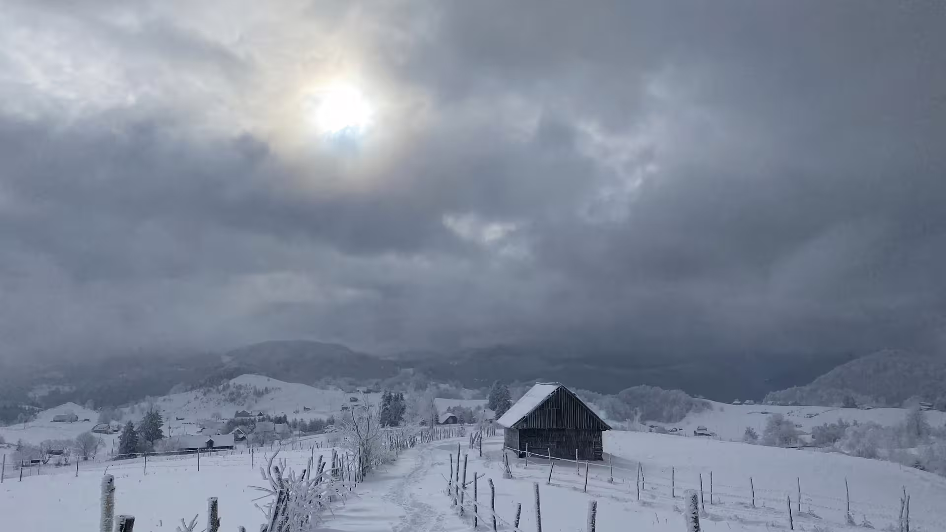 Snow-covered Transylvania village with wooden barn, fenced path, rolling hills, and dramatic winter sky