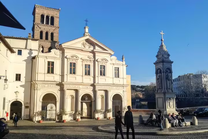 Visitors explore a sunlit Trastevere piazza with a historic Roman church and ornate cross monument in Rome.
