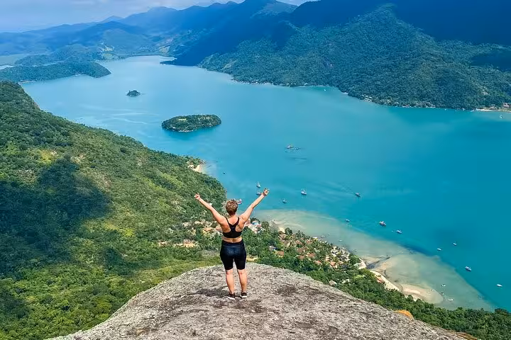 Traveler celebrates atop Peak Sugar Bread, overlooking stunning turquoise bay and lush greenery, perfect for adventure seekers.