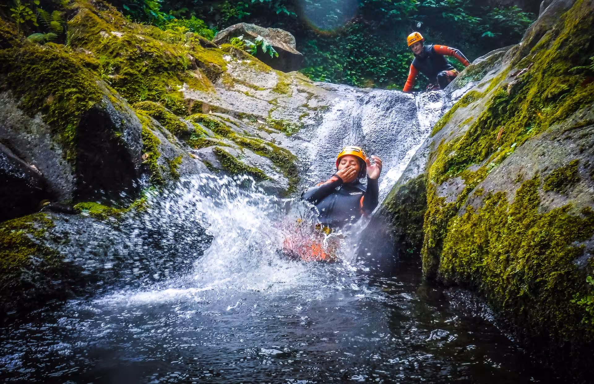 Traveler on Furnas canyoning tour sliding down a natural rock waterslide into a clear pool surrounded by green moss