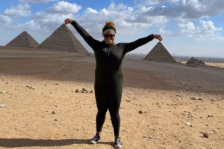 Traveler poses with arms outstretched in front of the iconic Giza Pyramids under a bright, cloudy sky.
