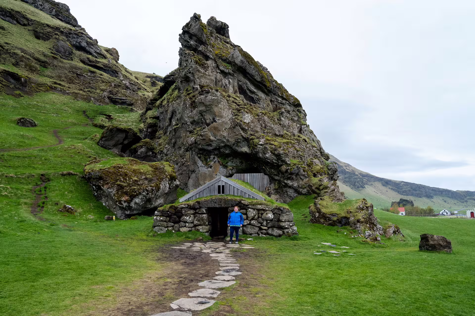 Traveler standing at historic turf house below dramatic rocky cliffs on Southeast Iceland Viking tour