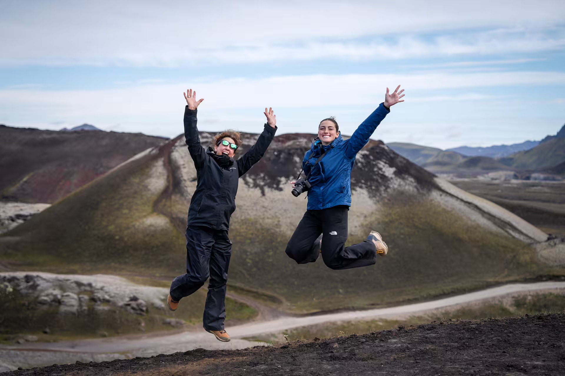 Two excited travelers jump joyfully in front of colorful volcanic highlands landscape on The Hidden Highlands Iceland adventure tour