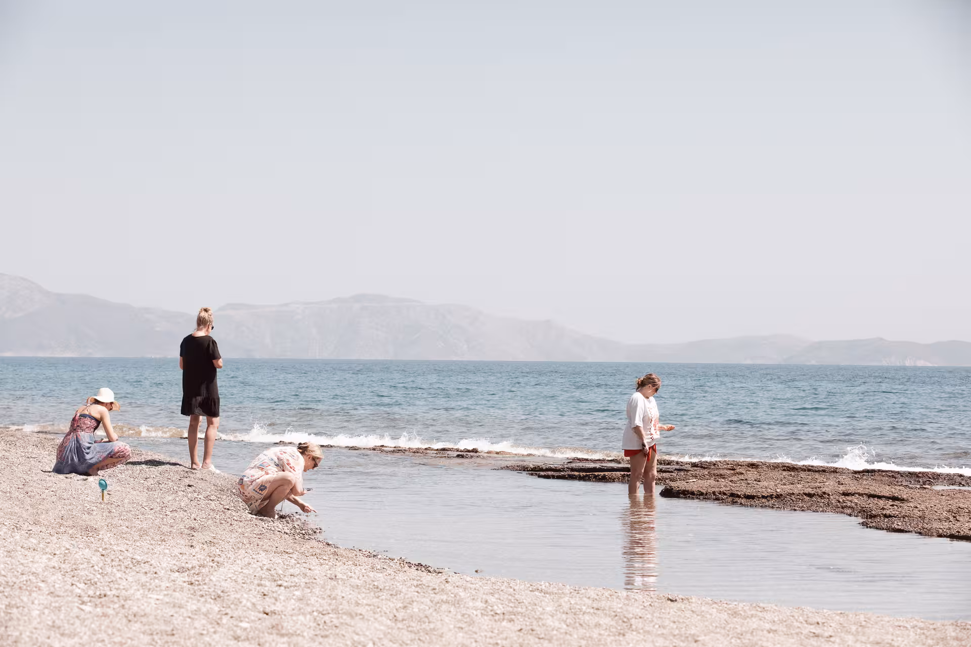 Travelers relax on a quiet Ikaria beach, paddling in shallow Therma waters with hazy Aegean mountains in the background