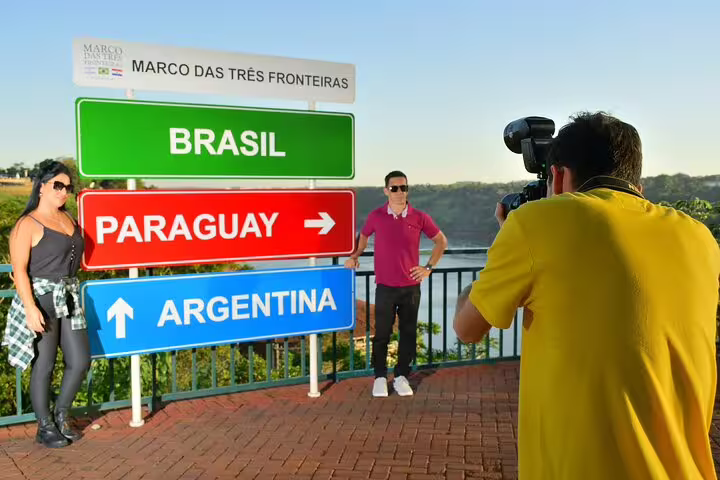 Visitors pose by Tres Fronteiras sign indicating directions to Brazil, Paraguay, and Argentina on a sunny day.