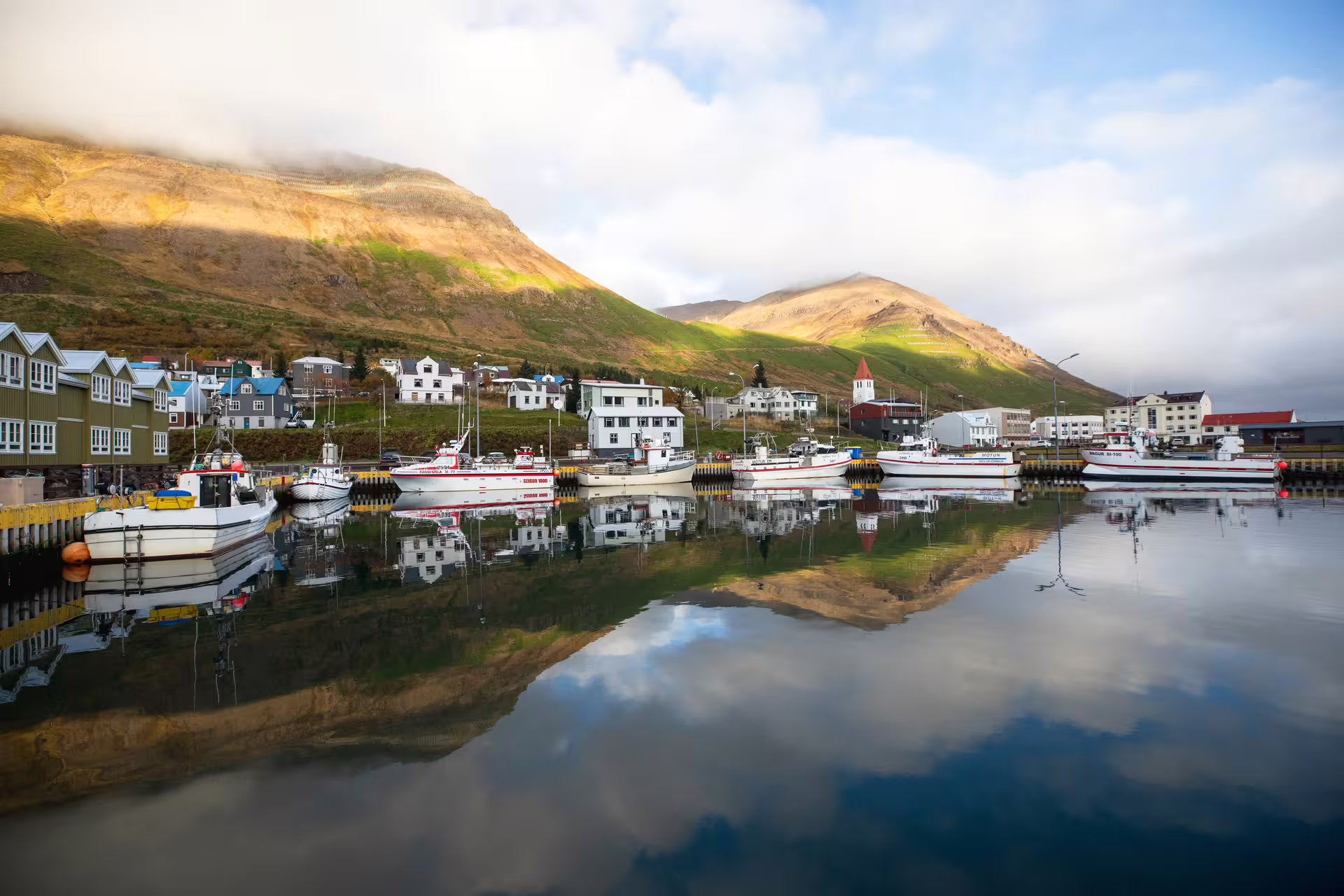 Fishing boats docked in calm Arctic fjord harbor with mountain reflections, Troll Peninsula Iceland