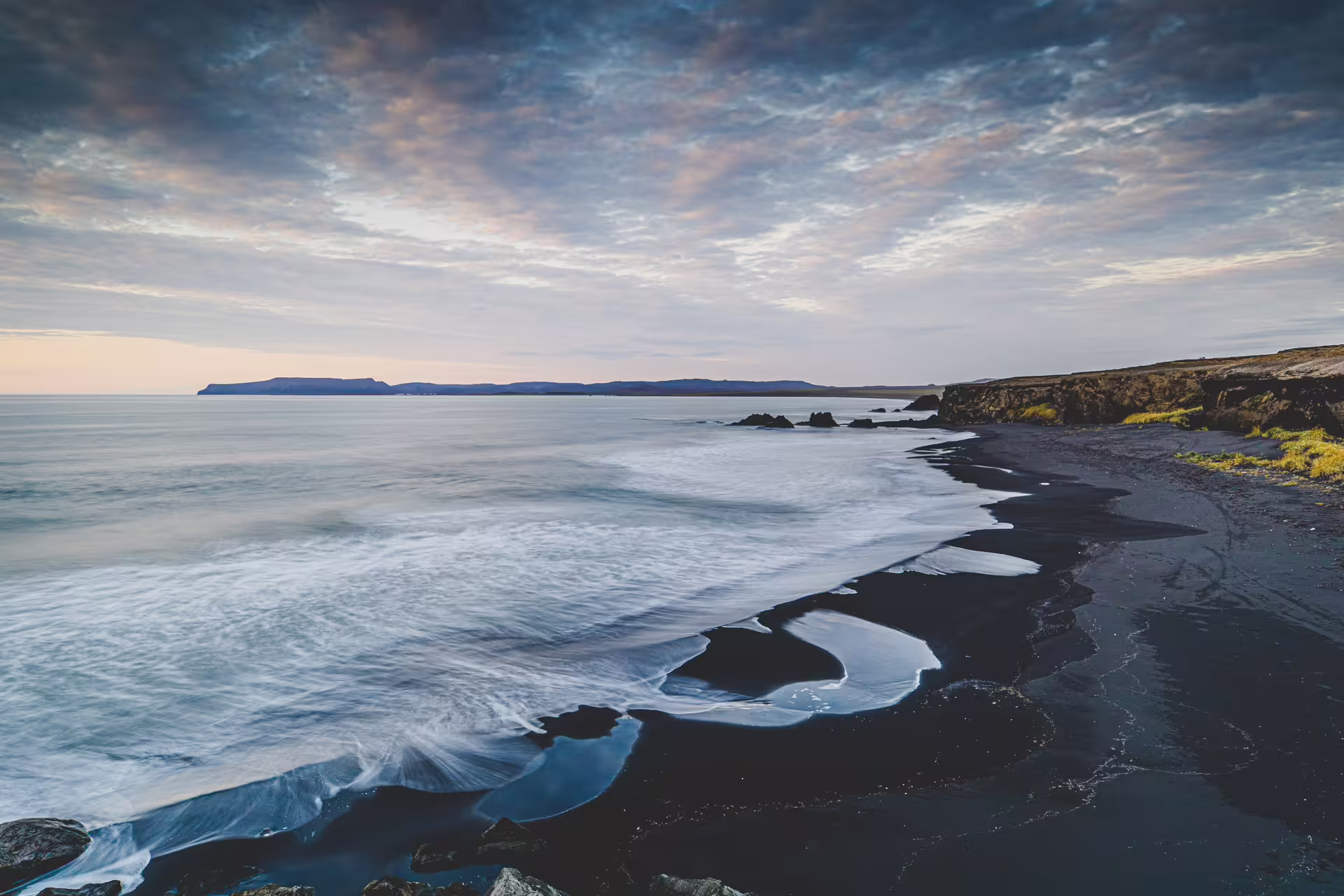 Black sand beach with gentle waves at dusk, rocky cliffs and distant mountains on Iceland's Troll Peninsula