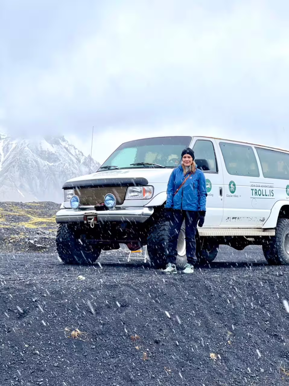 Woman in blue jacket standing by Troll.is super jeep during snowfall with snowy mountains in Iceland