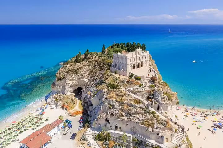 Aerial view of Santa Maria dell’Isola monastery on rocky promontory above crystal-clear Tropea beach in Calabria, Italy