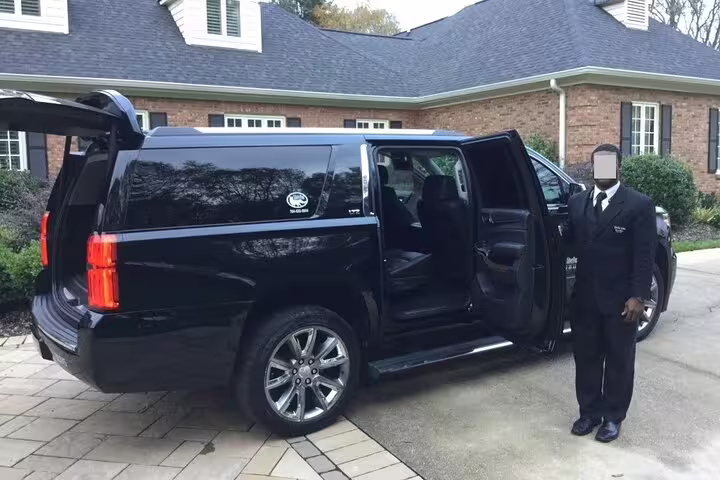 Professional chauffeur in black suit standing beside black Cadillac Escalade SUV with open door at residential home