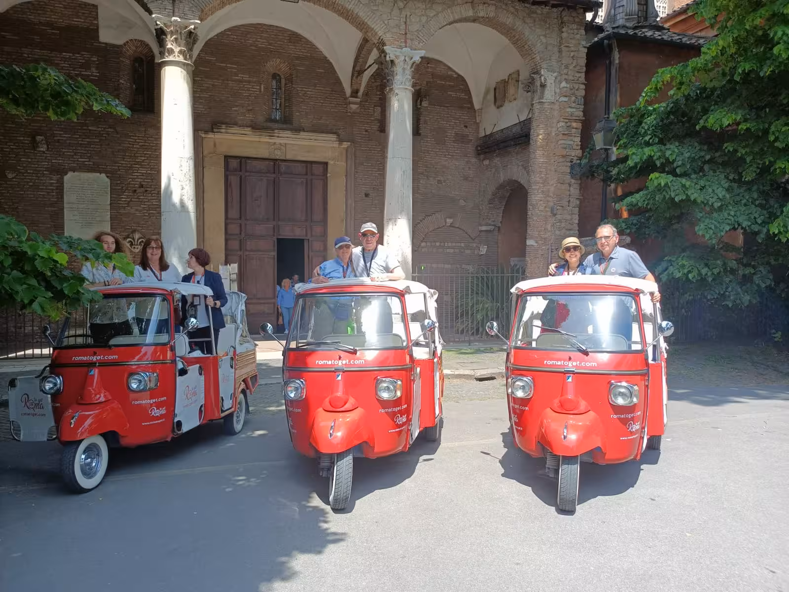 Group enjoying a tuk-tuk tour with red vehicles parked outside a historic Roman building, perfect for group adventures.