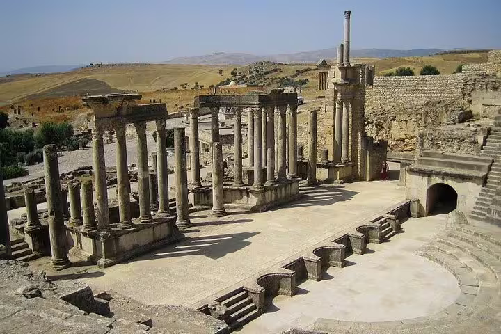 Ancient Roman ruins in Dougga, Tunisia, showcasing well-preserved columns and a theater under a clear blue sky.