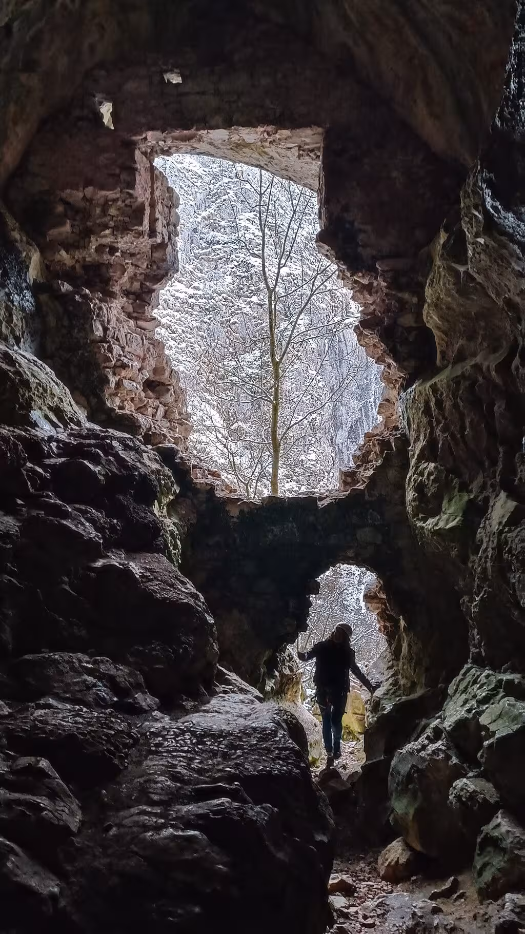 Hiker silhouetted at cave entrance with snowy trees visible through rocky opening in Turda Canyon