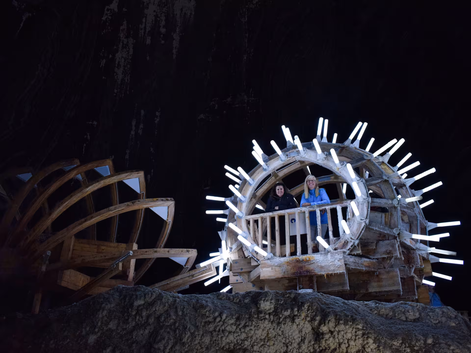 Two visitors standing inside illuminated wooden wheel structure at Turda Salt Mine underground cavern