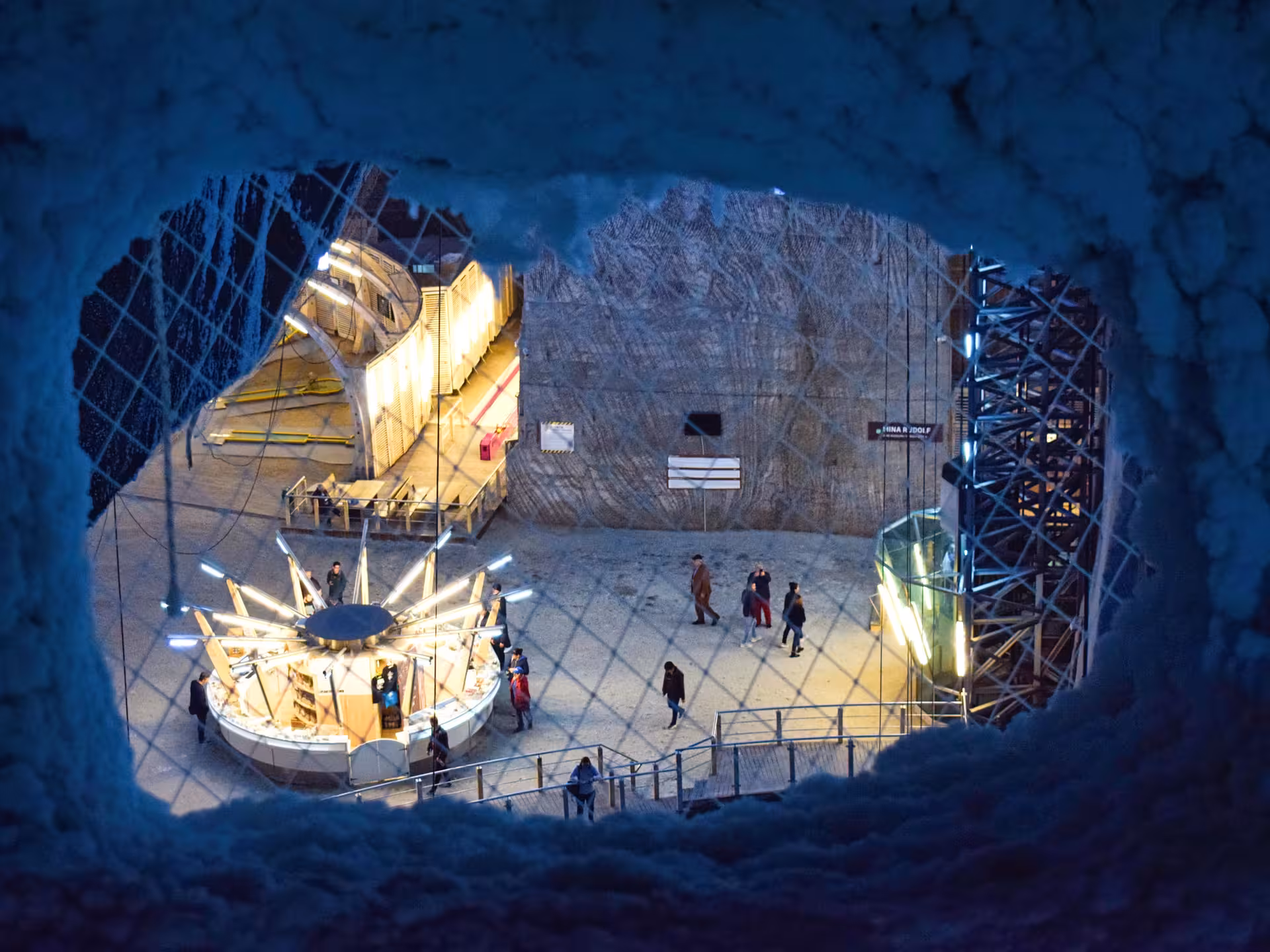 Aerial view through a salt cave opening at Turda Salt Mine showing illuminated underground attractions and visitors exploring the historic Rudolf mine chamber during a guided tour from Cluj