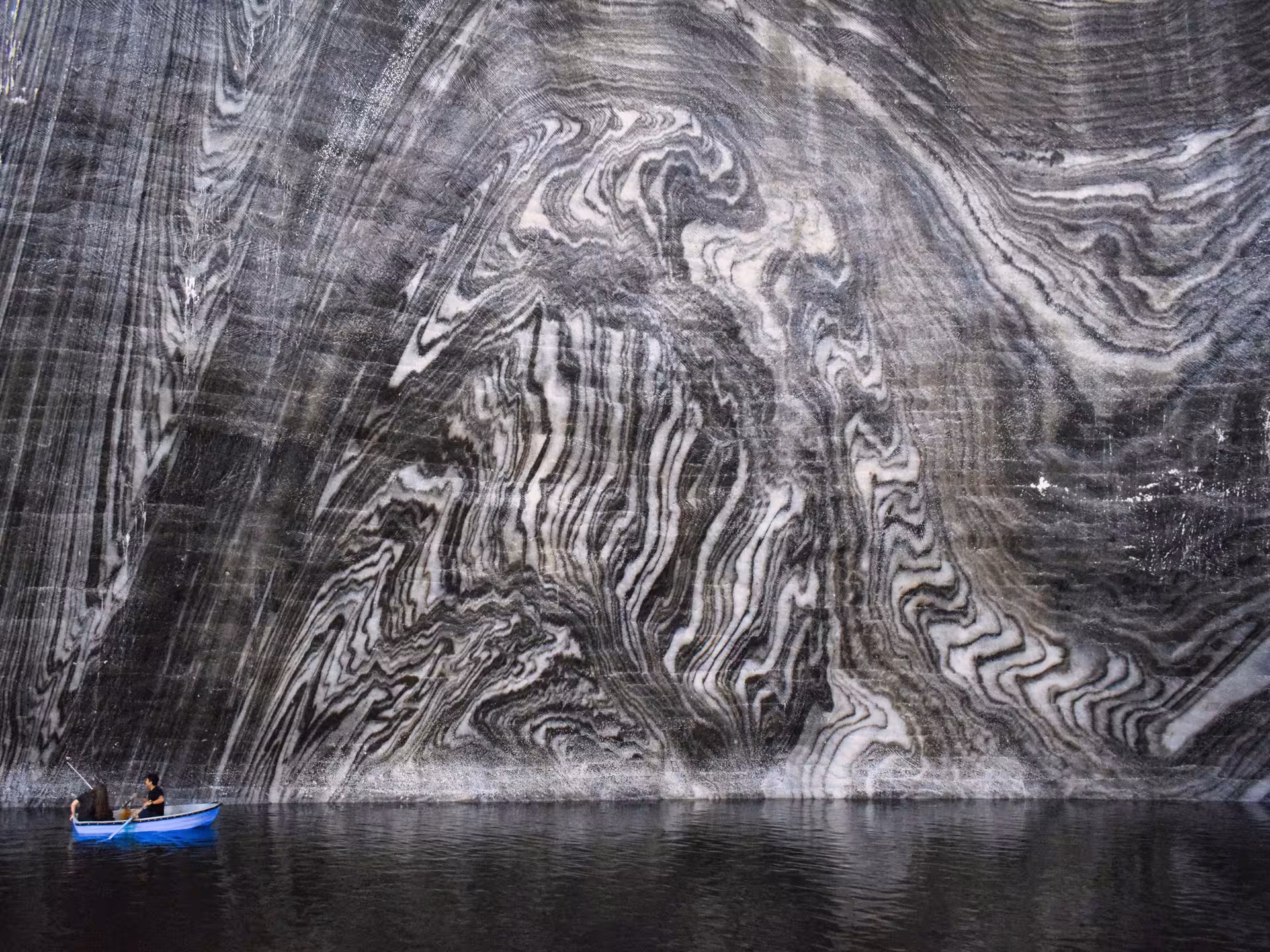 Two visitors rowing a blue boat on the underground lake inside Turda Salt Mine with swirling salt walls