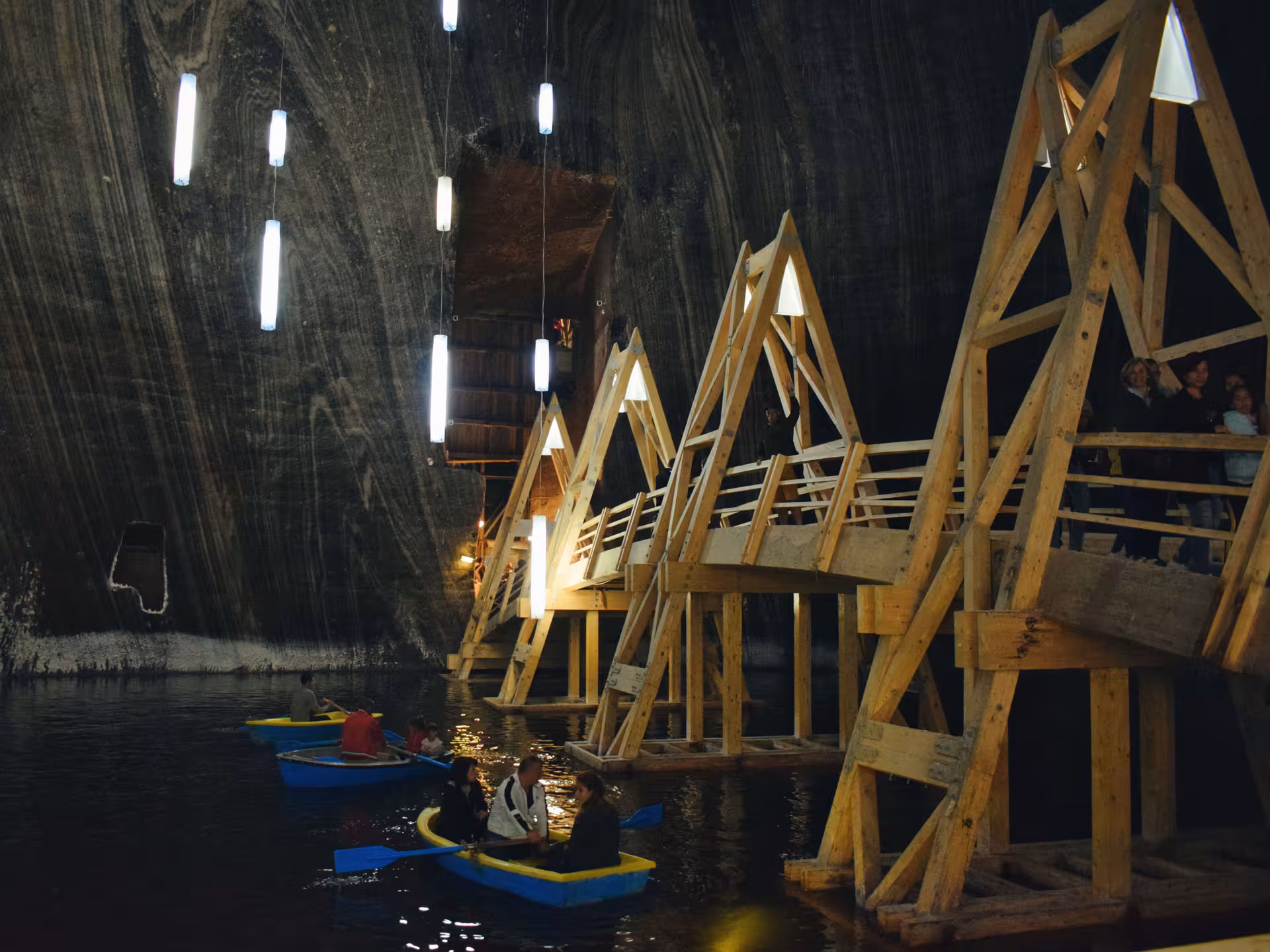 Visitors rowing blue boats on underground lake inside Turda Salt Mine with wooden walkways and hanging lights
