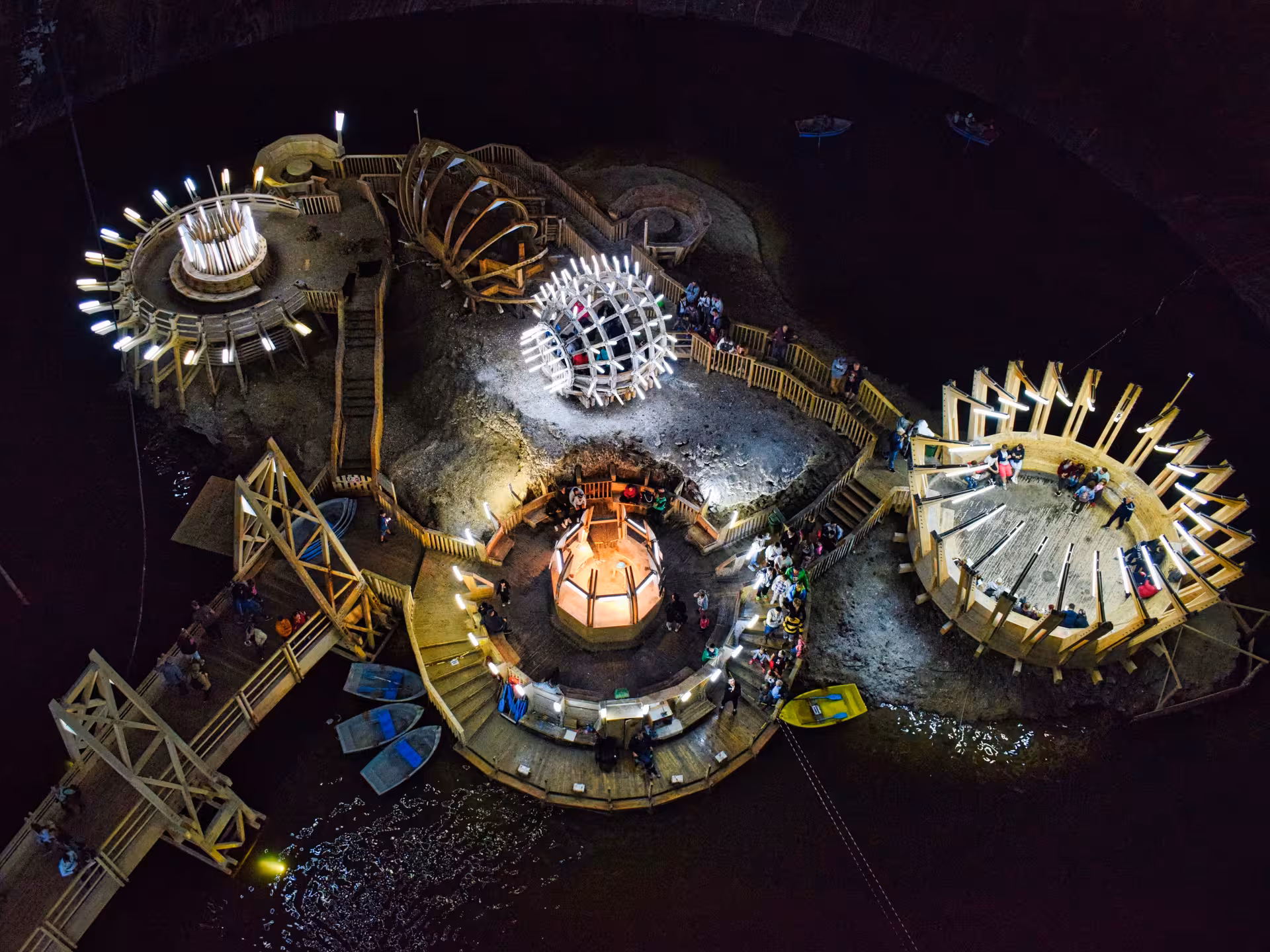 Aerial view of illuminated Turda Salt Mine interior with wooden structures, boats, and visitors exploring underground lake.