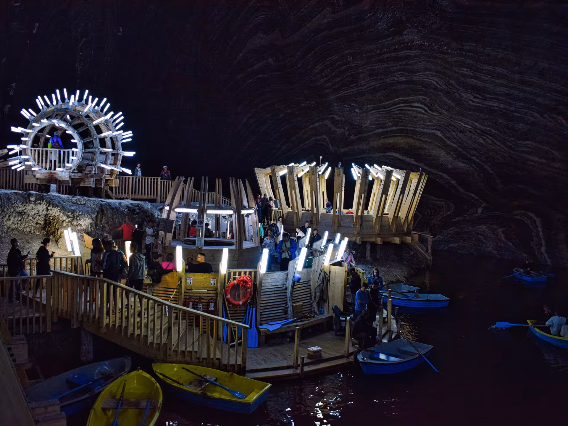 Tourists exploring illuminated wooden platforms and rowing boats on the underground lake at Turda Salt Mine Romania