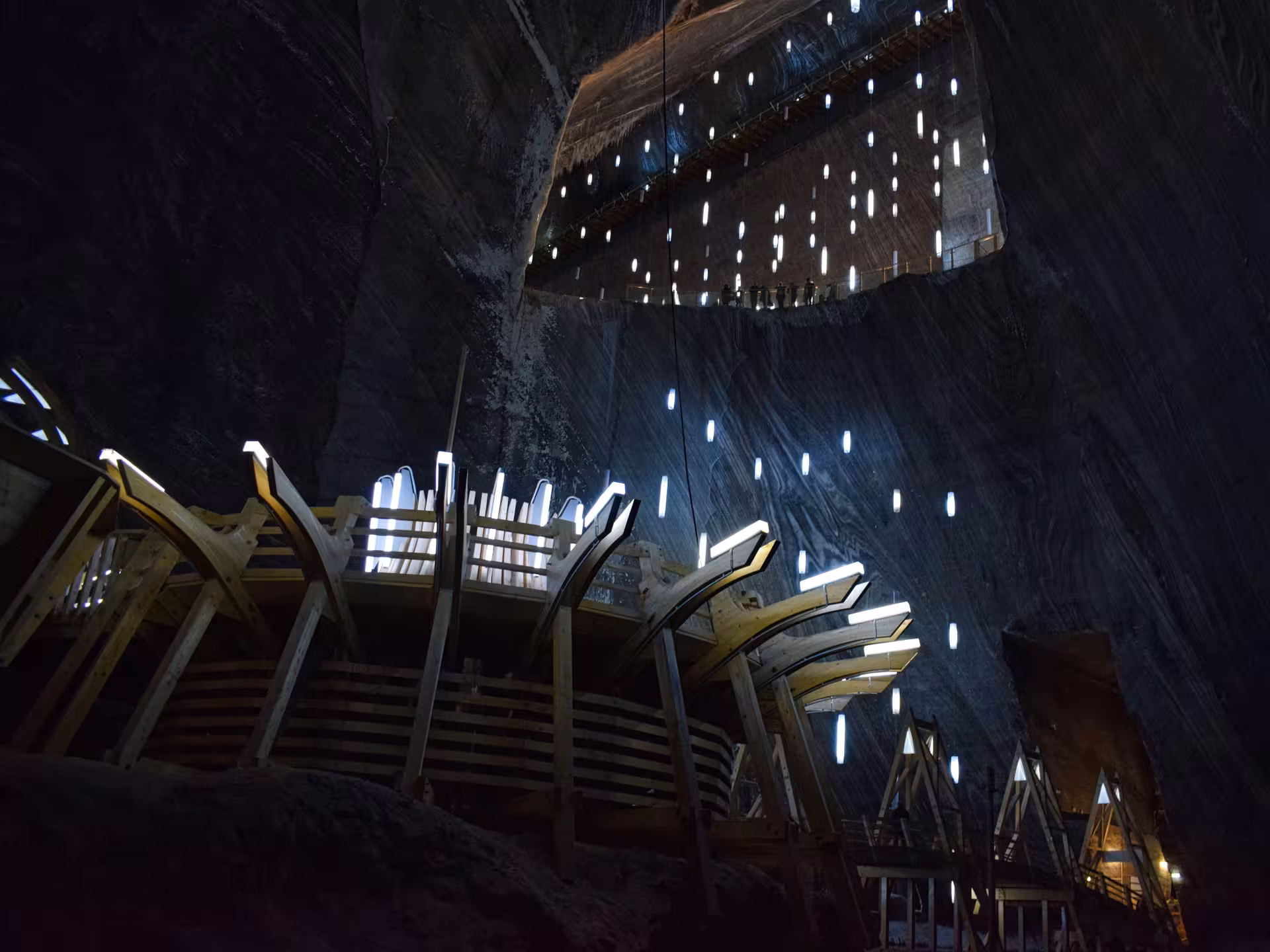 Spiral wooden staircase descending into Turda Salt Mine with glowing LED lights on dark salt walls