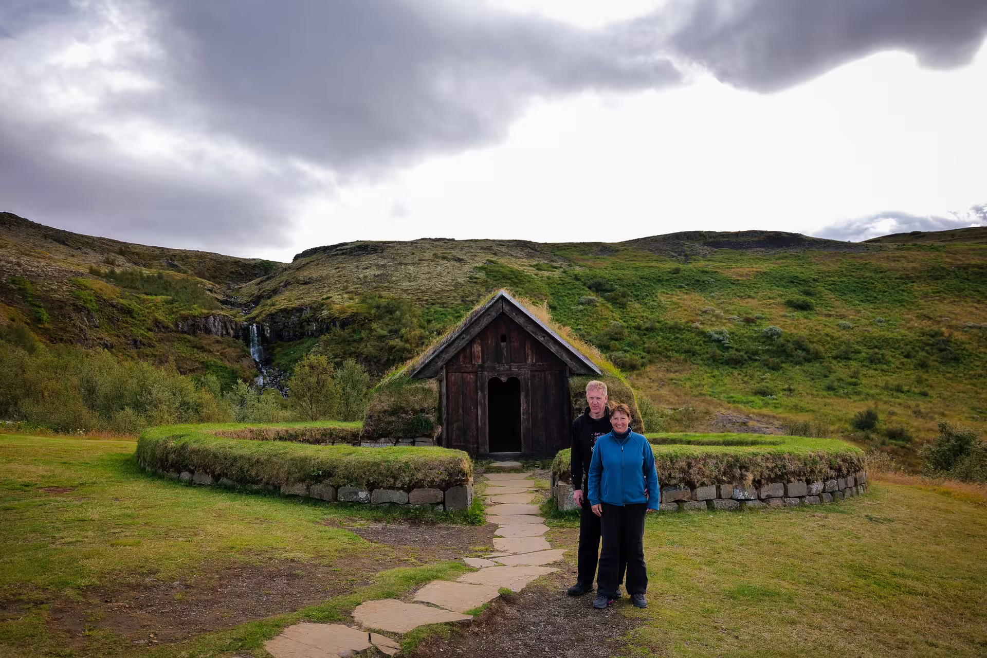 Couple posing before a historic turf-roofed church with waterfall and green Highland hills on The Hidden Highlands tour