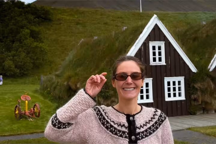 Smiling woman in Icelandic sweater and sunglasses in front of a traditional turf house in Westfjords