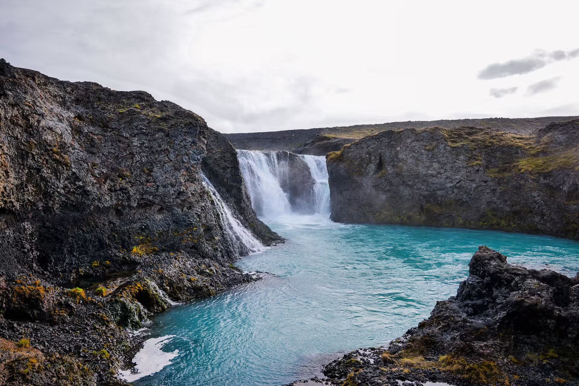 Stunning turquoise glacial river fed by powerful waterfall cascading over dark lava rock cliffs in Iceland highlands