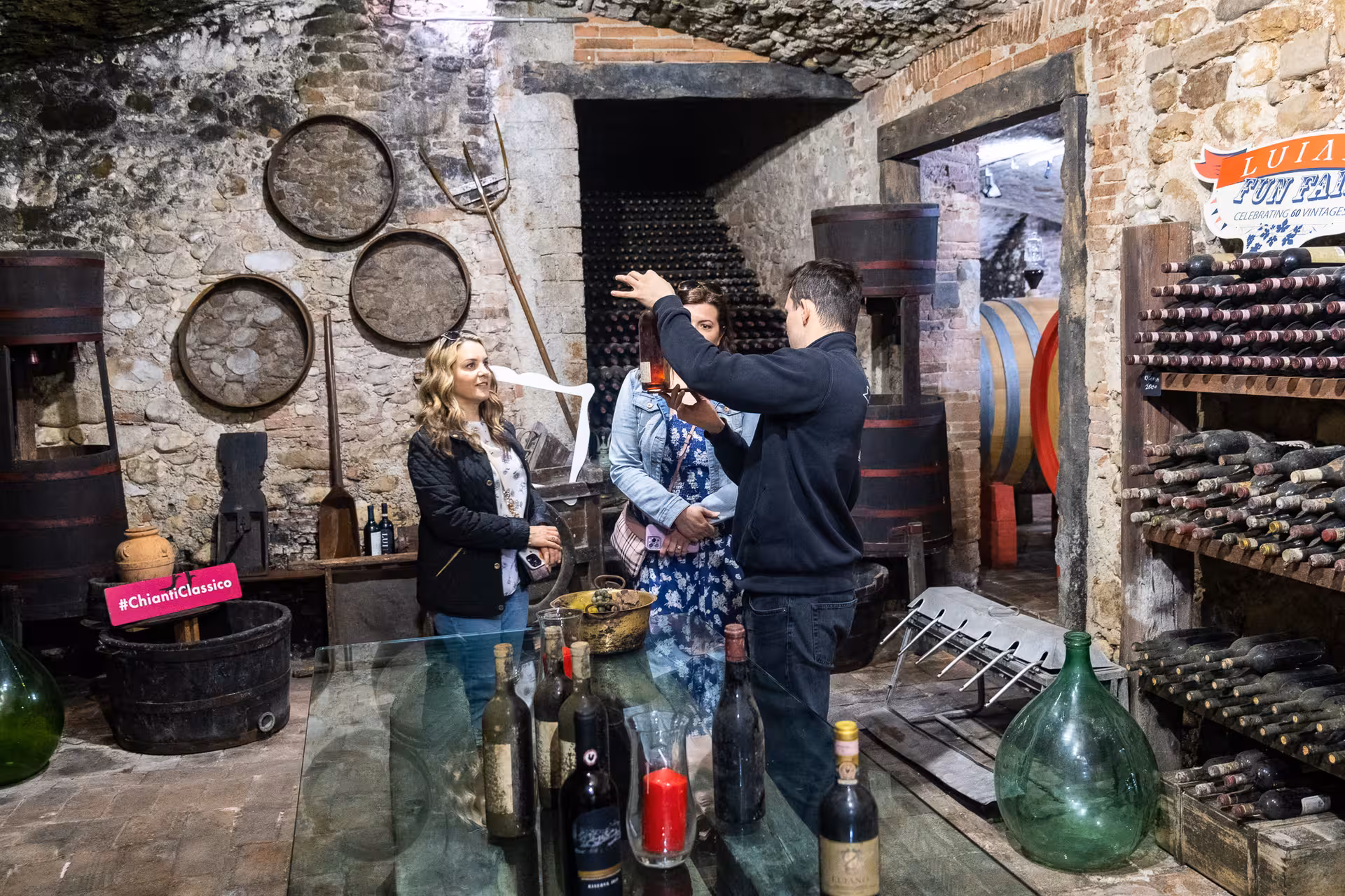 Tour guide showing wine bottles to visitors in a rustic Tuscan cellar during olive oil tasting tour.