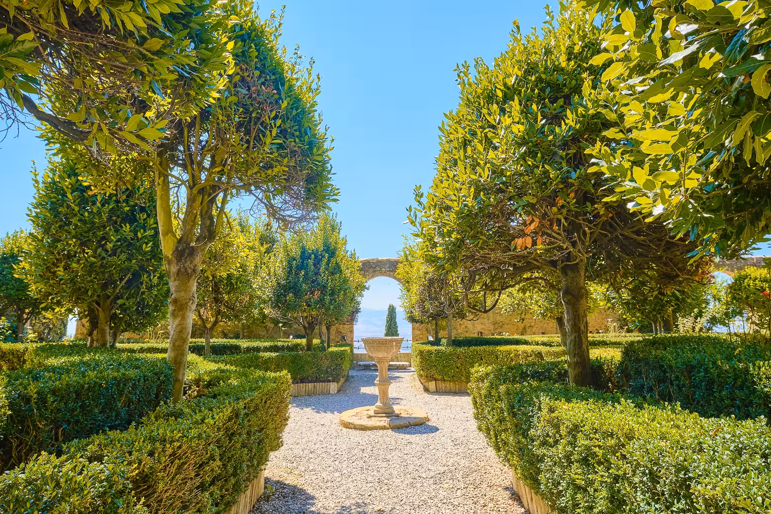 Stroll through a sunlit, manicured garden with lush greenery and a stone archway in Tuscany.