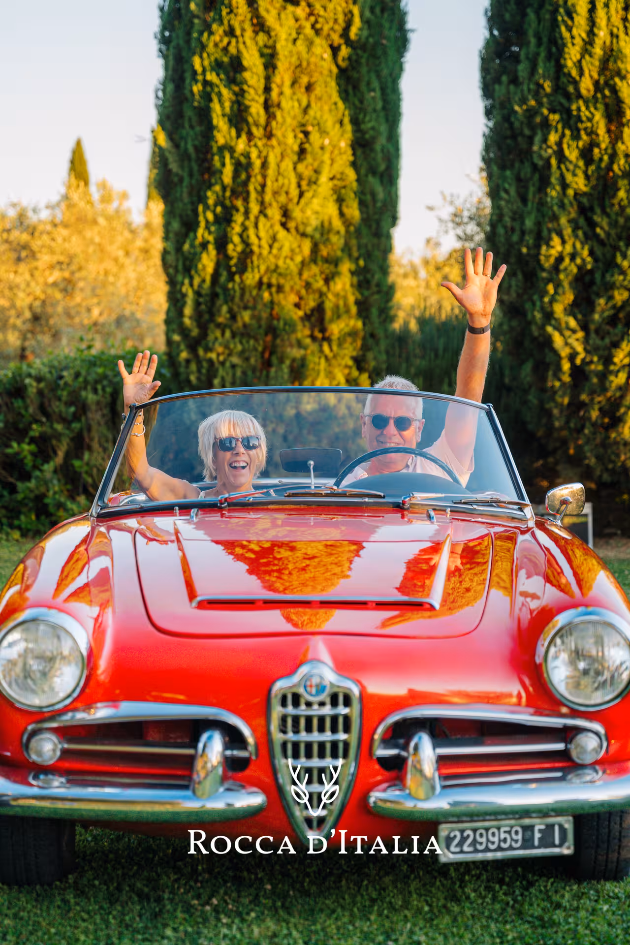 Two people joyfully ride in a classic red convertible amidst the beautiful Tuscan landscape.