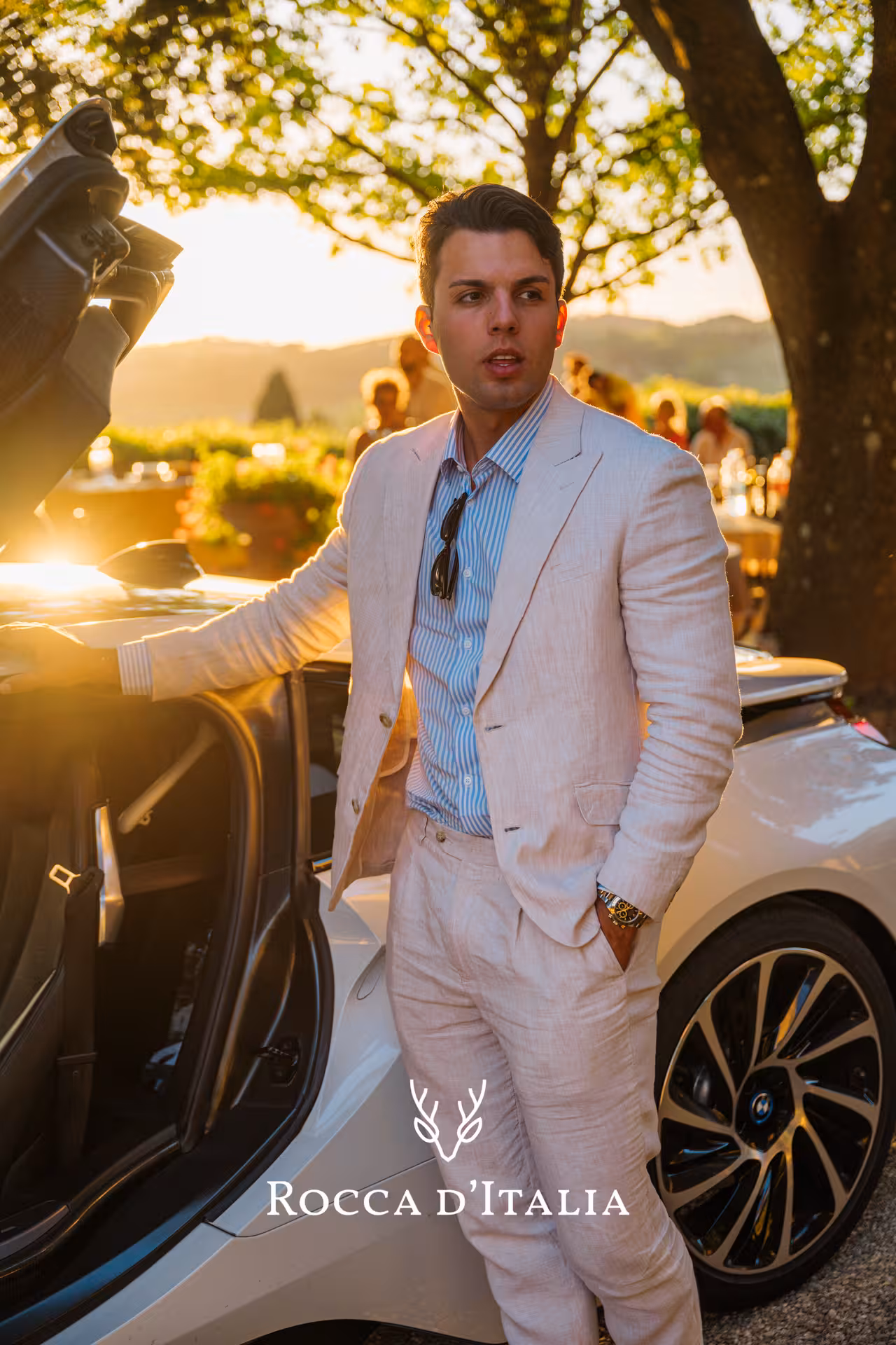 A person in a stylish suit stands by a supercar at sunset, capturing the elegance of the Tuscany Hills Experience.