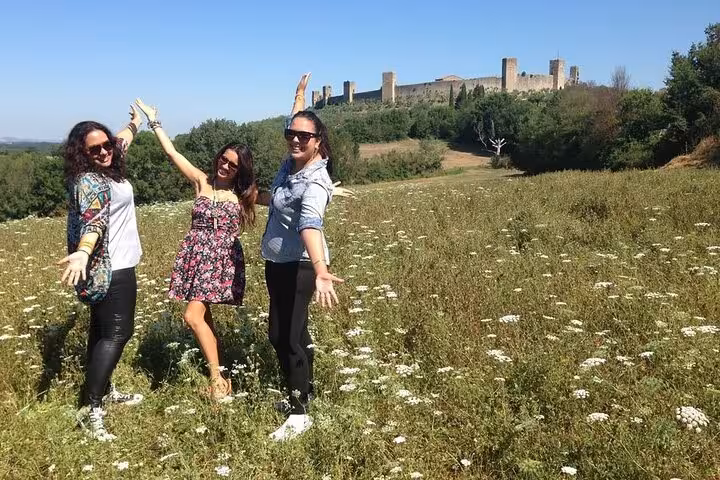Three happy tourists in a wildflower field pose with Tuscany's medieval fortress in the background under a clear sky.