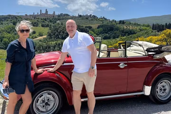 Couple enjoying a Tuscany tour with a vintage red Beetle car against scenic rolling hills backdrop.