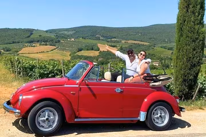 Tourists pointing at the Tuscan hills from a red vintage Beetle during a scenic drive.