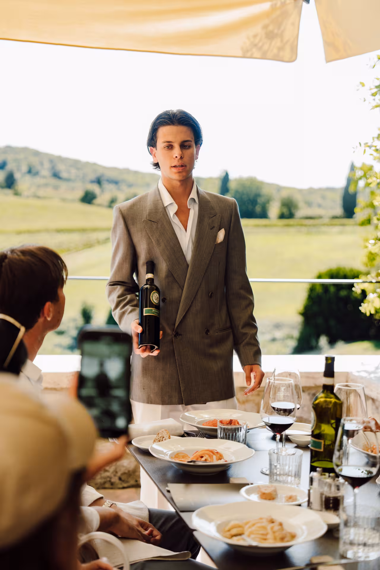 Sommelier presenting red wine bottle to guests at outdoor Tuscany dining terrace with vineyard views
